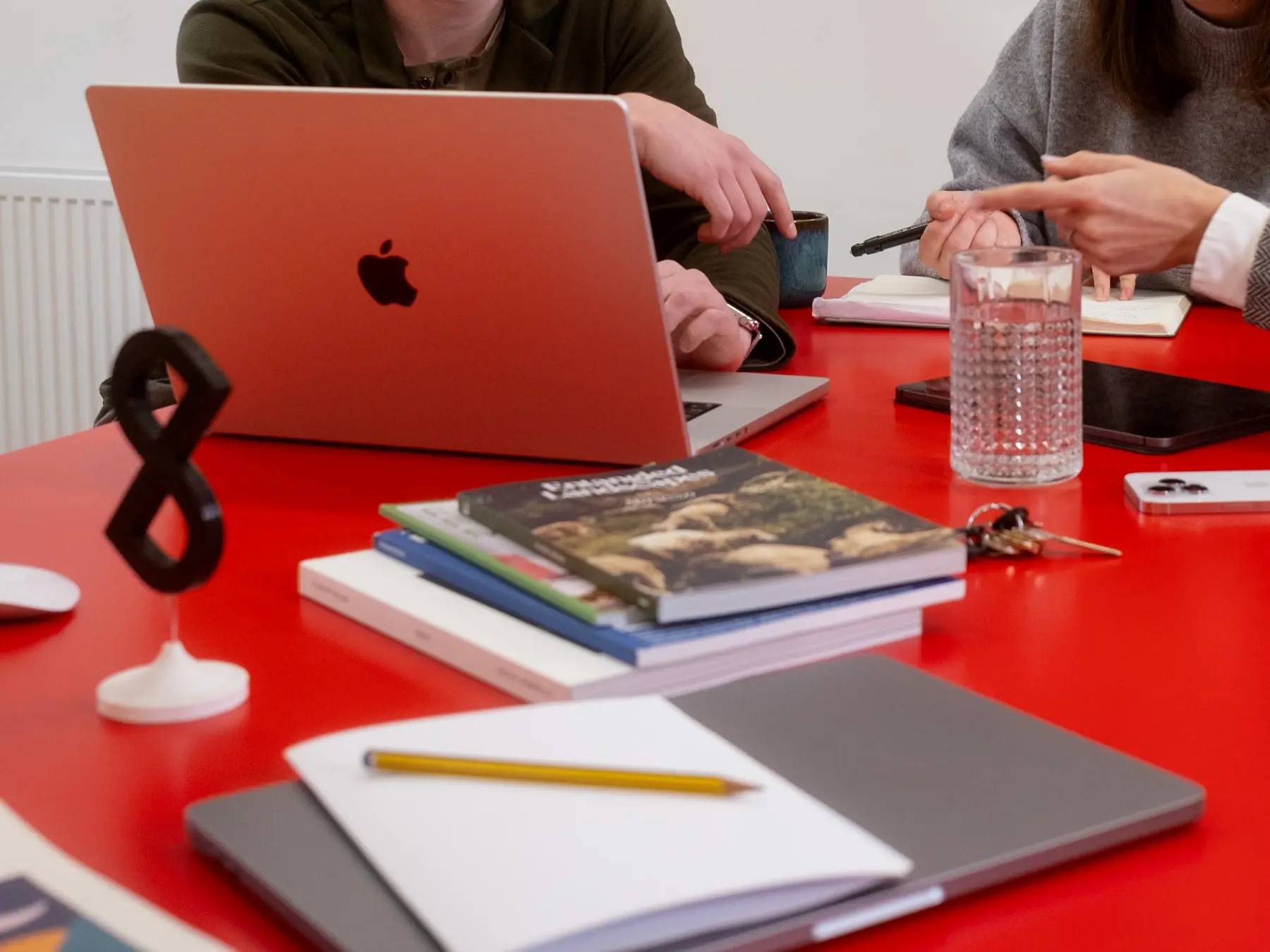 Two pagestrip designers discussing in front of a MacBook on a red table cluttered with magazines, pens, papers, and other everyday items.